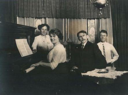 Old photo of mother father, adult daughter and teen son gathered around a piano.